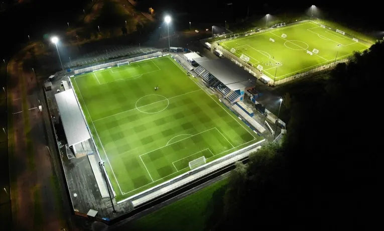 Toàn cảnh sân vận động Tameside của Curzon Ashton FC lung linh về đêm, cho thấy hệ thống chiếu sáng hiện đại.